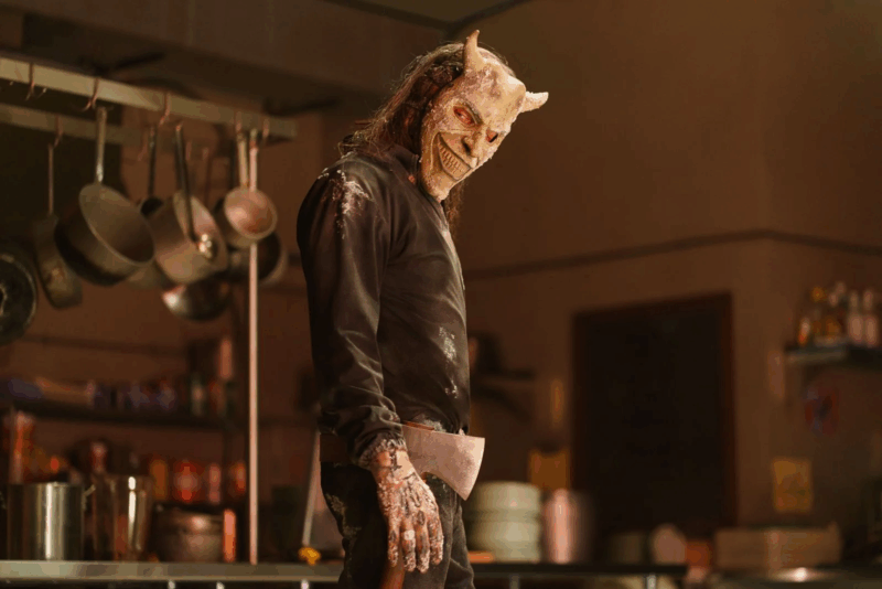 Um homem de cabelos longos e roupas escuras está em uma cozinha industrial iluminada por tons quentes. Ele veste uma máscara branca com chifres e expressão demoníaca, segurando uma faca grande em uma das mãos, com uma postura ameaçadora e olhar voltado para alguém fora do enquadramento.