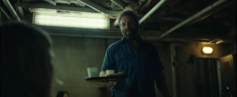 Cena do filme Animais PerigososNa imagem, Tucker oferece, com a mão direita, uma bandeja com dois copos com bebidas e um sanduíche para Zephyr, que está no canto esquerdo da imagem, apenas com a parte de trás da cabeça visível, em desfoque. Ao fundo, há uma mulher de cabelos escuros, apenas sua testa e o topo da cabeça estão visíveis. Tucker, no centro da imagem, usa uma camisa de botões na cor azul marinho. Ele é um homem branco, de cabelos e barba loiros, na faixa dos 40 anos de idade. O cenário se trata de um quartinho em um barco cheio de encanamentos. 