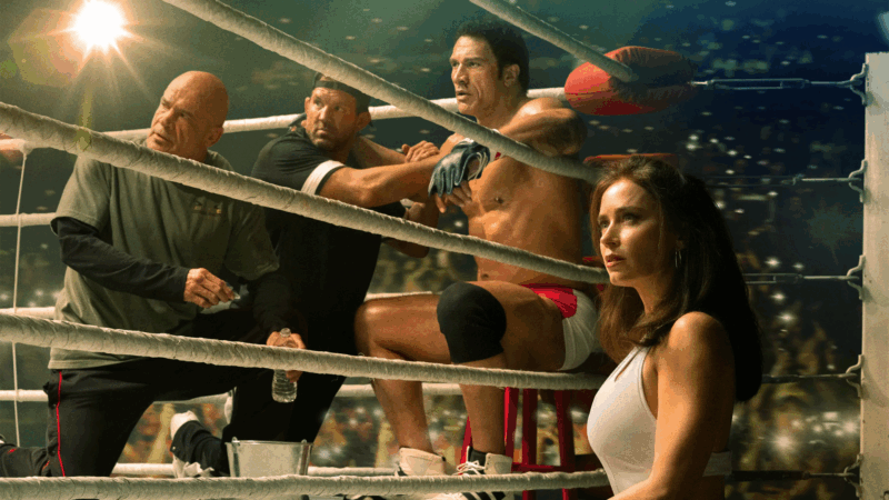 Uma cena do filme Coração de Lutador, vista de dentro de um ringue de luta, focada em quatro personagens em um momento de alta tensão. Da esquerda para a direita: Bas Rutten, como um treinador, é um homem mais velho, careca, vestindo uma camiseta verde-escura. Ele se inclina para a frente com uma expressão de intensa preocupação e seriedade, olhando para o lutador. Atrás do lutador, Ryan Bader o segura firmemente pelos ombros. Ele está vestido de preto e seu rosto também mostra foco e preocupação, como se estivesse tentando conter ou dar apoio. No centro da imagem, sentado em um banco vermelho no corner do ringue, está o lutador Mark Kerr, interpretado por Dwayne Johnson. Ele está sem camisa, exibindo um físico musculoso, e veste shorts de luta brancos com detalhes em vermelho e uma joelheira preta. Sua expressão é de angústia e exaustão; seus olhos estão arregalados e fixos, e sua boca está entreaberta. À direita, do lado de fora das cordas, está Dawn, interpretada por Emily Blunt. Com cabelos longos e escuros e vestindo uma regata branca, ela olha para a cena com uma expressão de profunda tristeza, angústia e preocupação.