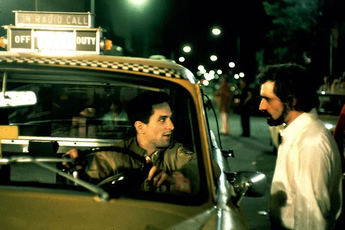 Fotografia colorida mostra Martin Scorsese e Robert De Niro em um momento de interação no set de Taxi Driver (1976). Scorsese, com barba e óculos escuros, veste camisa branca e calça escura, enquanto De Niro, caracterizado como Travis Bickle, usa jaqueta militar e camisa xadrez. Ao fundo, é visível uma rua urbana com prédios típicos de Nova York, refletindo o ambiente da década de 1970.