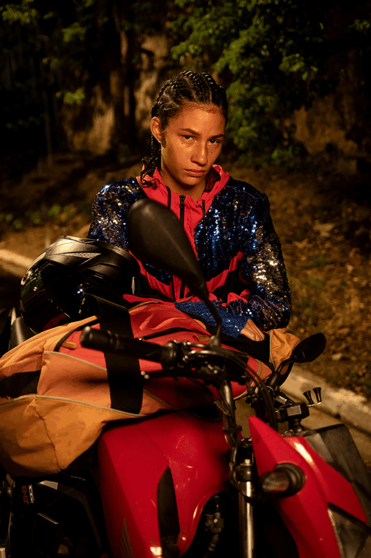 Na imagem, há uma mulher negra de tranças. Ela está vestindo um casaco colorido e está em cima de uma moto. No seu colo, há uma mala grande na cor laranja. Em seu rosto, há lágrimas escorrendo e expressões que remetem a tristeza. 