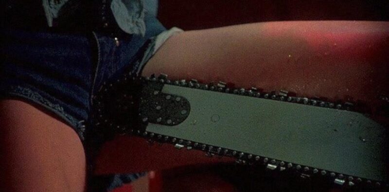 Cena do filme O Massacre da Serra Elétrica 2Na imagem, no canto direito, uma serra elétrica prateada encosta na região da genital de uma mulher, que está no canto esquerdo.. A parte íntima está protegida, coberta por um short jeans curto que ela usa. A pele da mulher é na cor clara e as cores da imagem são escuras, mas com a presença de tons vermelhos. Da personagem, apenas vemos o short e as coxas, que estão abertas, em posição de penetração. 