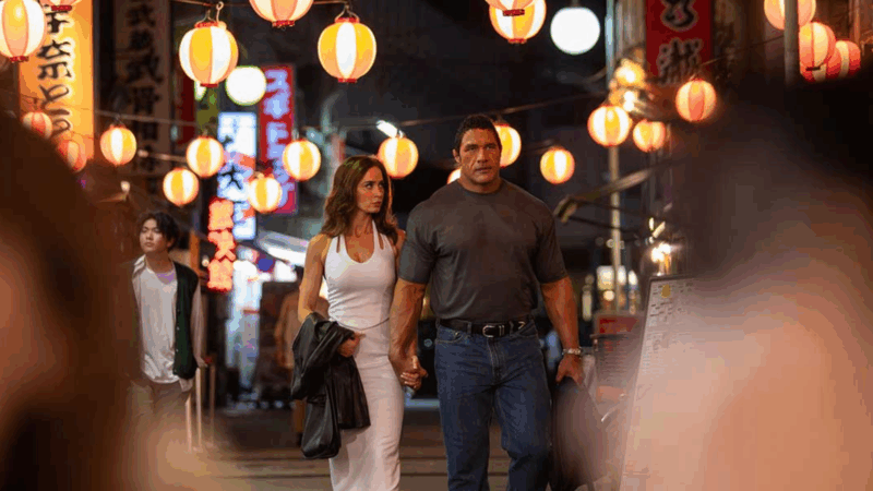 Uma cena noturna do filme Coração de Lutador que mostra Dwayne Johnson e Emily Blunt caminhando juntos por uma rua movimentada no Japão. O casal está de mãos dadas no centro da imagem, ambos com expressões sérias e focadas. Dwayne Johnson, à direita, veste uma camiseta cinza-escura justa que acentua seu físico extremamente musculoso, combinada com calça jeans. Emily Blunt, à esquerda, usa um vestido branco longo e justo e segura uma jaqueta escura em seu braço. O cenário é o destaque da imagem: a rua está iluminada por dezenas de lanternas de papel redondas, com listras brancas e alaranjadas, penduradas em fios sobre suas cabeças, criando uma iluminação quente e atmosférica. Ao fundo, letreiros verticais com caracteres japoneses e outras luzes urbanas complementam a cena vibrante. A imagem é capturada de uma perspectiva que inclui elementos desfocados em primeiro plano, dando a sensação de que estamos observando o casal de perto.