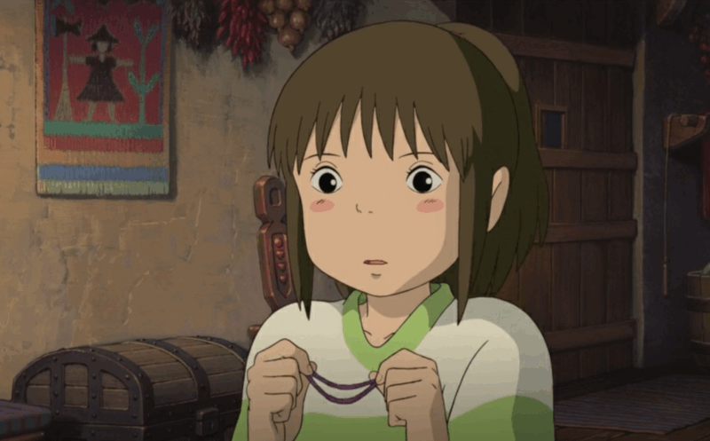 Cena da animação A viagem de chihiro, focando na protagonista. Ela é uma menina de cabelos castanhos e olhos grandes, com uma expressão de surpresa e bochechas coradas. Ela veste uma blusa branca e verde e segura um elástico de cabelo roxo. Ao fundo, na parede, há um pequeno quadro ou tapeçaria colorida com a figura de uma bruxa de chapéu pontudo e vestido escuro, além de um baú e outros elementos rústicos do cenário.