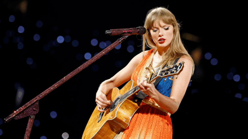 Taylor Swift se apresentando no palco durante a The Eras Tour. Ela está cantando com uma expressão concentrada enquanto toca um violão acústico de madeira clara. A cantora veste um vestido laranja cintilante e sem mangas, com lábios pintados de vermelho e seu característico delineado gatinho. Ao seu lado, um pedestal e um microfone estão cobertos de glitter avermelhado. O fundo é escuro e desfocado, pontilhado por luzes azuis e brancas, sugerindo a imensidão da plateia em um estádio.