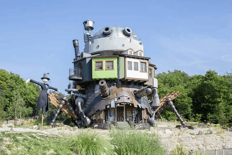 Esta é uma fotografia da réplica em tamanho real do Castelo Animado, do filme "O Castelo Animado" do Studio Ghibli, localizada no Ghibli Park no Japão. A estrutura é uma construção fantástica e assimétrica, parecendo uma colagem de diferentes casas, canos e peças de metal, com uma base escura e enferrujada que se assemelha a uma criatura com pernas e canhões. Em um nível intermediário, há paredes coloridas, uma verde e outra branca. O cenário é um parque com vegetação rasteira, rochas e uma densa floresta ao fundo, sob um céu azul claro. À esquerda, está o personagem Espantalho, com um chapéu-coco e um casaco esfarrapado.