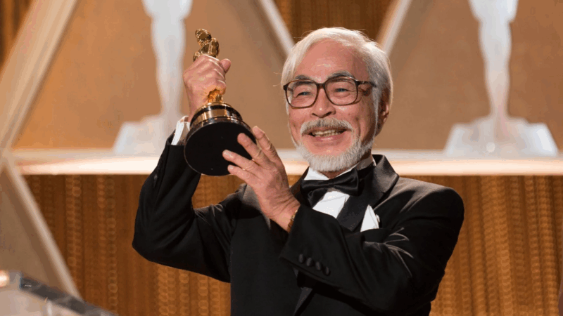 Esta é uma fotografia de Hayao Miyazaki, o aclamado diretor do Studio Ghibli. Ele é um homem idoso, de cabelos e barba brancos, e usa óculos de armação escura. Ele veste um smoking preto com uma gravata borboleta e sorri calorosamente para a câmera. Com as duas mãos, ele segura orgulhosamente uma estatueta do Oscar, que é dourada e brilhante. O fundo é predominantemente dourado e desfocado, sugerindo que ele está em um palco durante uma cerimônia de premiação.
