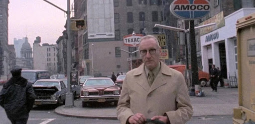  Cena do documentário Nova ’78. William S. Burroughs, um homem branco, idoso, de sobretudo claro e óculos, caminha por uma rua de Nova York na década de 1970, cercado por carros antigos e placas de postos de gasolina.