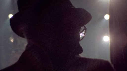 Cena do documentário Nova ’78. William S. Burroughs, de costas, é visto em silhueta, de chapéu e óculos, com as luzes do palco ao fundo criando um contraste entre sombra e brilho