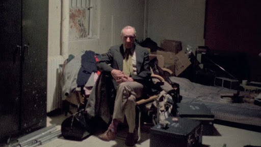 Cena do documentário Nova ’78. William S. Burroughs, um homem branco, idoso, de estatura média, sentado em uma cadeira no canto de um quarto simples e desorganizado, cercado por roupas, caixas e equipamentos.