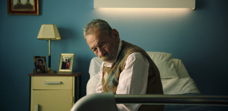 Na fotografia, um homem branco idoso de barba grisalha está sentado à beira de uma cama em um quarto de hospital com paredes azuis. Ele usa um colete de tricô, majoritariamente da cor verde oliva, sobre uma camisa social branca enquanto olha para baixo, com expressão melancólica. Ao fundo, há uma luminária acesa e uma mesa de cabeceira amarela com porta-retratos e um abajur.
