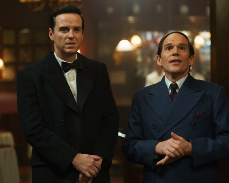 Cena do filme Blue Moon. Na imagem, há dois homens brancos. O da esquerda veste um terno preto com gravata borboleta da mesma cor. O da direita usa um terno azul com gravata vinho.