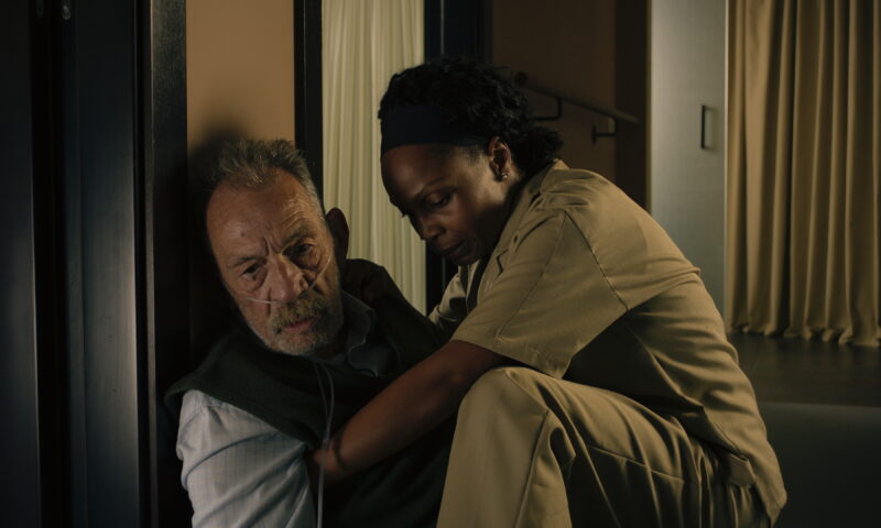 No frame, um homem branco idoso com tubos de oxigênio no nariz está sendo amparado no chão por uma mulher negra vestida em um uniforme totalmente bege. Ela o segura com firmeza e preocupação, enquanto ele apresenta um semblante de exaustão. A cena ocorre em um ambiente interno de iluminação suave, com cortinas claras e paredes escuras ao fundo.