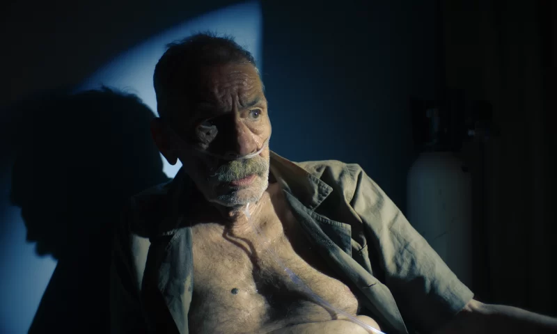 A imagem mostra um homem branco idoso magro, com expressão de espanto, sentado em um ambiente escuro, iluminado por um foco de luz branco em meio a uma parede azulada. Ele está com a camisa desabotoada, com tubos de oxigênio no nariz, e parece fragilizado. Um cilindro de oxigênio branco aparece parcialmente ao fundo, reforçando o clima de vulnerabilidade.