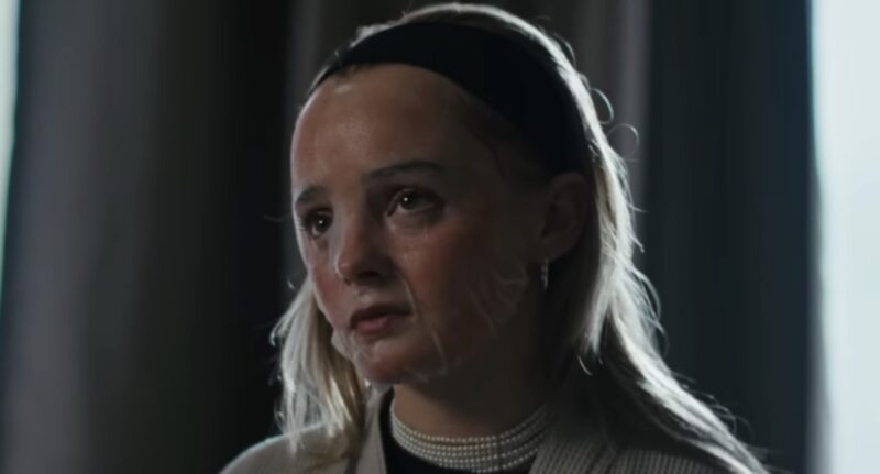 Cena do filme mostra, uma adolescente branca de cabelo loiro segurado por uma faixa preta enquanto usa uma máscara transparente na pele, que está avermelhada. Seu olhar é melancólico e desanimado. Ao fundo, tons azulados são evidentes em meio as cortinas. 