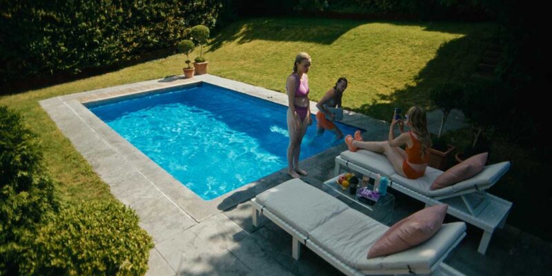 Cena do filme Babystar. Três pessoas estão à beira de uma piscina azul, que está rodeada por uma área verde. Luca, uma adolescente branca e loira, está em pé, olhando para a mãe, uma mulher branca e loira, que está sentada na cadeira de praia olhando para o celular, ambas estão de biquíni, rosa e laranja, respectivamente. O pai, homem branco e loiro, está usando um calção laranja e segura um laptop.