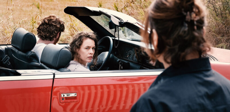 Cena de Mirrors No.3. Uma mulher de cabelo cacheado e suéter claro está no banco de passageiro de um carro conversível vermelho, olhando para trás, na direção de uma pessoa cujas costas e cabelo preso estão em primeiro plano. Um homem com cabelo cacheado está sentado no banco do motorista ao lado dela.