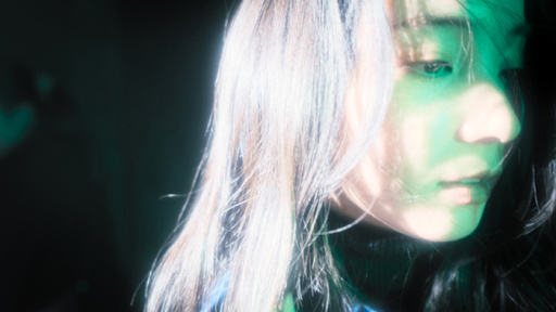 O rosto de uma jovem é parcialmente iluminado por uma luz verde-azulada que incide lateralmente, criando um contraste suave entre brilho e sombra. Seus cabelos longos caem sobre o rosto, e o olhar baixo sugere introspecção ou melancolia. O fundo é escuro, reforçando a atmosfera etérea e contemplativa da cena.