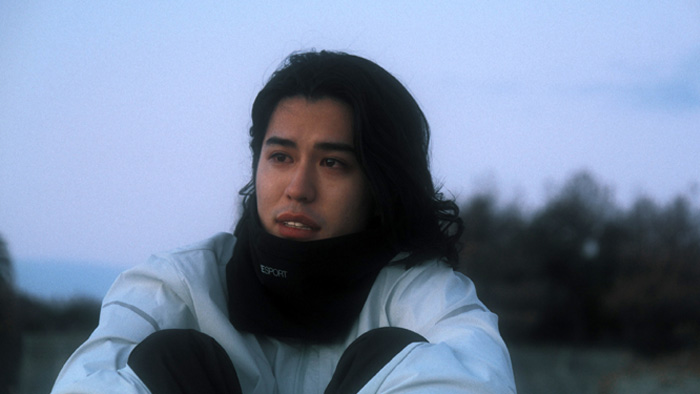 Um jovem está sentado ao ar livre, envolto em um casaco branco e cachecol preto, olhando para frente com expressão de tristeza contida. A luz natural do amanhecer ou entardecer banha suavemente seu rosto, enquanto o fundo desfocado revela árvores e um céu pálido, sugerindo um momento de solidão e reflexão.