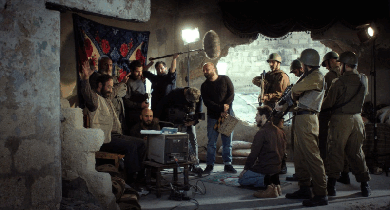 Fotografia de um grupo de homens reunidos em uma sala em ruínas, com paredes em tons claros descascadas e iluminação branca artificial e intensa. À esquerda, um homem sentado ergue as mãos, enquanto uma câmera e um microfone gravam em meio à técnicos de filmagem que o cercam. Soldados armados, à direita, completam a cena, em que a linha entre encenação e realidade parece se dissolver.