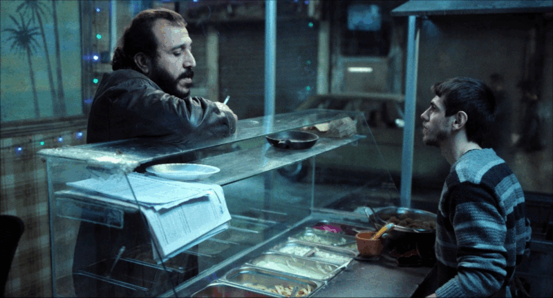 Foto de dois homens conversando, separados por um balcão de comida, sob uma luz fria e azulada. Um deles, apoiado no vidro, fuma enquanto fala e o outro observa em silêncio, sentado diante de pratos e utensílios. A cena carrega uma sensação de cansaço cotidiano, marcada pelo reflexo do vidro e pela penumbra do ambiente.