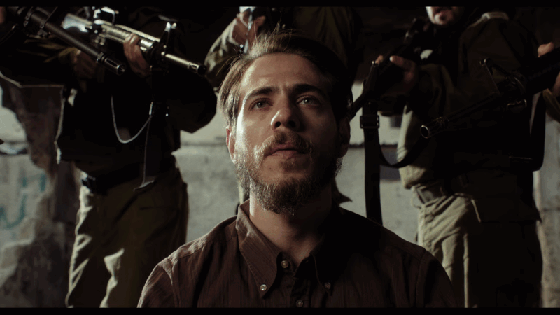 Fotografia de um homem de expressão séria e olhar distante ocupa o centro do enquadramento em um ambiente sombrio e deteriorado iluminado por uma luz branca que destaca seu rosto. Atrás dele, figuras uniformizadas empunham armas, compondo uma cena tensa que mistura fragilidade e ameaça. O contraste entre a serenidade do homem e a iminência da violência cria uma atmosfera carregada de dramaticidade, onde a linha entre ficção e realidade parece prestes a se romper.