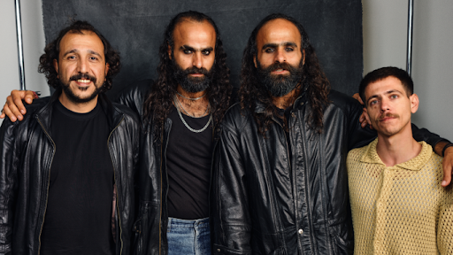 Diante de um fundo neutro, a foto destaca quatro homens posando lado a lado, com expressões que alternam entre a confiança e a introspecção. Vestidos em roupas escuras, sobretudo jaquetas de couro, e exibindo cabelos longos e barbas marcantes, eles formam um grupo cuja presença impõe identidade e coesão. A proximidade física e os olhares diretos à câmera revelam cumplicidade e força coletiva, sugerindo tanto um vínculo pessoal quanto uma postura artística compartilhada.