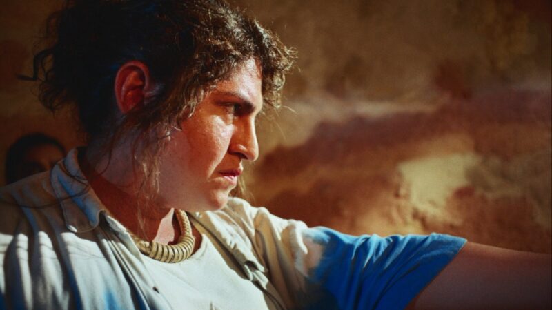 Cena do filme Morte e Vida Madalena Na imagem, Madalena aponta uma arma para sua frente, o objeto não está enquadrado na cena. A personagem está com o braço esquerdo estendido, usa uma camisa de botões que está aberta, na cor azul em degradê com o branco. Veste uma peça branca por baixo e um colar grosso com discos enfileirados que parece ser feito do material sisal. Madalena é travesti e possui cabelos cacheados na cor castanha, que estão presos. O fundo é um cenário de set de filmagens, uma parede rochosa na cor marrom. 