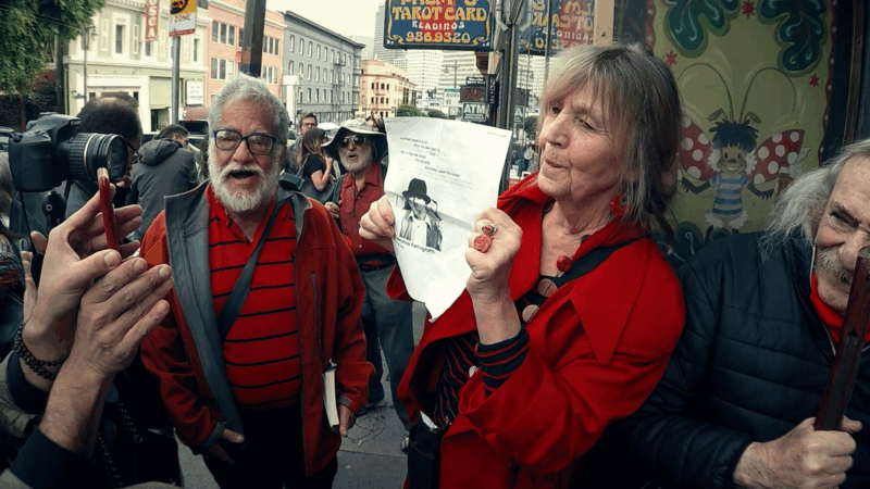 Numa cena de rua do documentário 'A Última Batida', um grupo de pessoas se reúne na calçada. Em primeiro plano, uma mulher idosa de casaco vermelho segura um folheto com a foto de um homem, enquanto um homem idoso de barba branca, óculos e colete vermelho gesticula ao seu lado. Ao fundo, outros membros do grupo, prédios da cidade e uma placa de 'Tarot Card Reading' são visíveis.