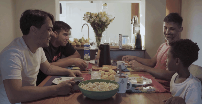 Quatro pessoas estão sentadas em volta de uma mesa de café da manhã. Ao lado esquerdo, há um homem com camiseta branca e um jovem adulto de camiseta preta. Do lado oposto da mesa, há um menino com camiseta preta e um homem de camiseta salmão. Todos estão com expressões felizes e encaram a criança mais nova, sentada à ponta direita. Em cima da mesa há uma bacia de pipoca, pão de queijo, garrafa de café, caixa de leite, pratos e canecas. 