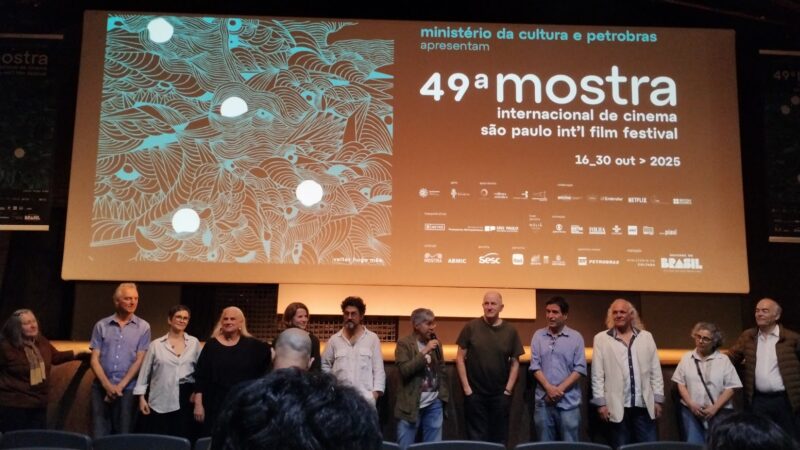 Um grupo numeroso de integrantes da equipe e do elenco está alinhado, com Lina Chamie no centro, à frente de uma tela de cinema que exibe a arte oficial da 49ª Mostra Internacional de Cinema de São Paulo. Lina Chamie segura um microfone enquanto os outros a assistem com atenção.