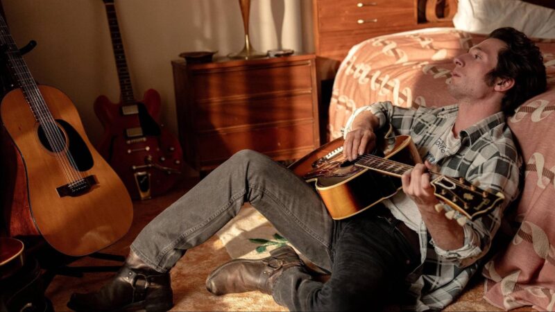 Homem branco com cabelos escuros, sentado no chão apoiado na cama com um violão de frente para uma janela