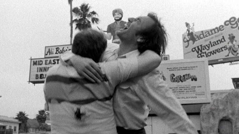 Cena e preto e branco do filme O Estado das Coisas. Dois homens se abraçam no meio da rua. Um deles está um buraco de tiro nas costas, com sangue na camisa. Já o outro, está gritando assustado com o disparo. Ao fundo, estão outdoors e letreiros de estabelecimentos, além de palmeiras.