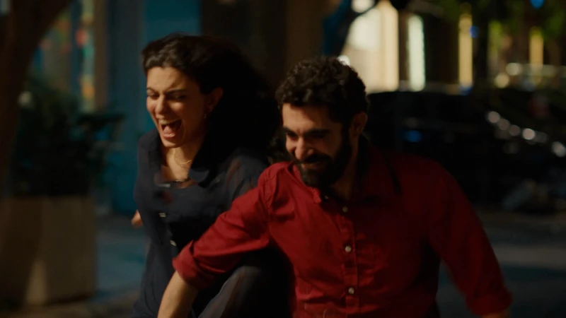 Cena do filme Um Mundo Triste e Belo (2025). À noite, o casal corre de mãos dadas por uma rua iluminada, rindo juntos. A mulher, de cabelos escuros e soltos, usa uma blusa azul escura. O homem, de barba e cabelo cacheado, veste uma camisa vermelha. As luzes da cidade e o desfoque do fundo sugerem liberdade e alegria espontânea