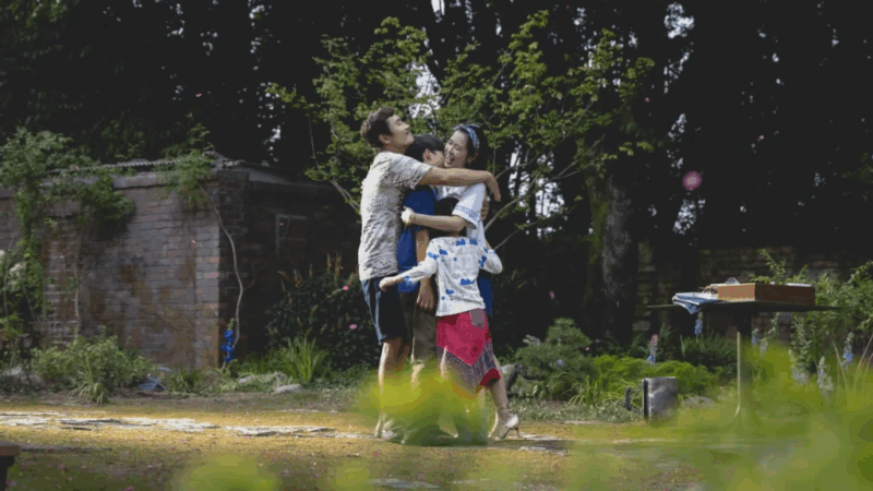 Ao ar livre, um homem, uma mulher e duas crianças se abraçam em meio a um jardim verdejante e iluminado pelo sol. O gesto é espontâneo e afetuoso, transmitindo alegria e leveza. No fundo, há árvores altas e um muro de tijolos coberto por vegetação, compondo um cenário de harmonia familiar.