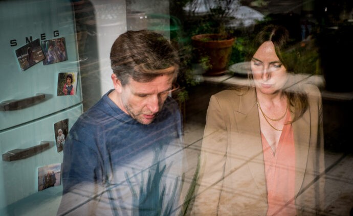 Cena do filme Maturidade. Na imagem, da esquerda para a direita, é possível ver um homem branco e de cabelo castanho vestindo uma camisa de mangas longas na cor azul. Ao seu lado, há uma mulher branca de cabelos castanhos, vestindo uma blusa laranja e um casaco marrom. Ao fundo, é possível ver uma geladeira azul com fotografias coladas. A foto foi tirada através de um vidro, sendo possível ver o reflexo do exterior na imagem.