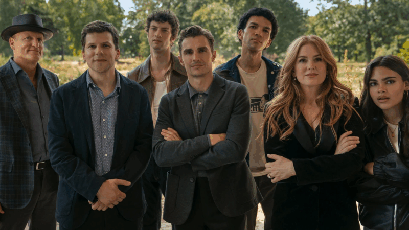 Cena de Truque de Mestre - O 3° Ato. A cena mostra um grupo diverso de oito pessoas posam lado a lado, encarando diretamente a câmera com expressões que variam entre sorrisos discretos e seriedade. A composição frontal e simétrica destaca a sensação de união e mistério, reforçada pelo personagem central de braços cruzados. O fundo desfocado de vegetação e a luz natural suave valorizam as cores e os rostos, enquanto as roupas — que vão do formal ao casual — e a diversidade entre os retratados adicionam complexidade. O resultado é uma imagem contemporânea, com tom dramático e enigmático.