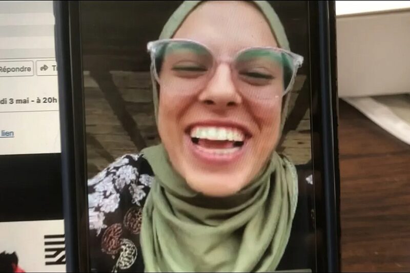 Cena do filme Guarde o Coração na Palma da Mão e Caminhe.Na imagem, há um celular em chamada de vídeo com Fatima Hassouna. O celular está apoiado em uma mesa de madeira e atrás dele há uma outra tela, logada em um e-mail. Hassouna é uma mulher palestina, na faixa dos 25 anos. Ela usa óculos arredondados, o lenço de sua cultura chamado de hijab, na cor verde, e uma roupa estampada. Ela está sorrindo. 