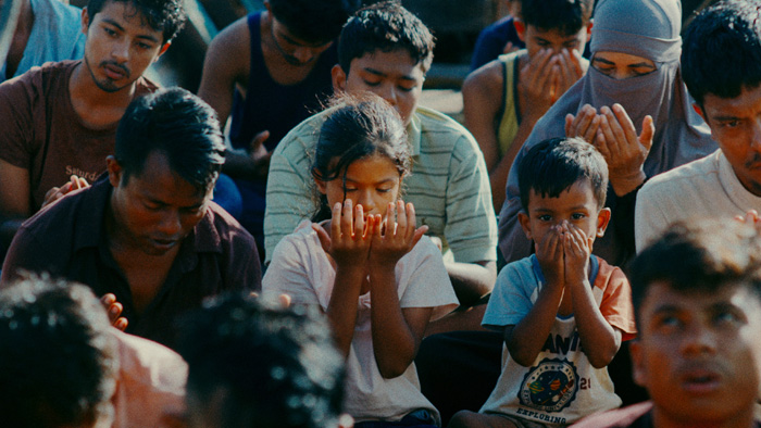 Cena do filme Terra Perdida. Somira, com uma camiseta branca e Shafi, vestindo uma camiseta estampada, ajoelhados entre outros refugiados, participam de uma oração coletiva em uma embarcação. Sérios, estão com as mãos juntas e erguidas rezando.