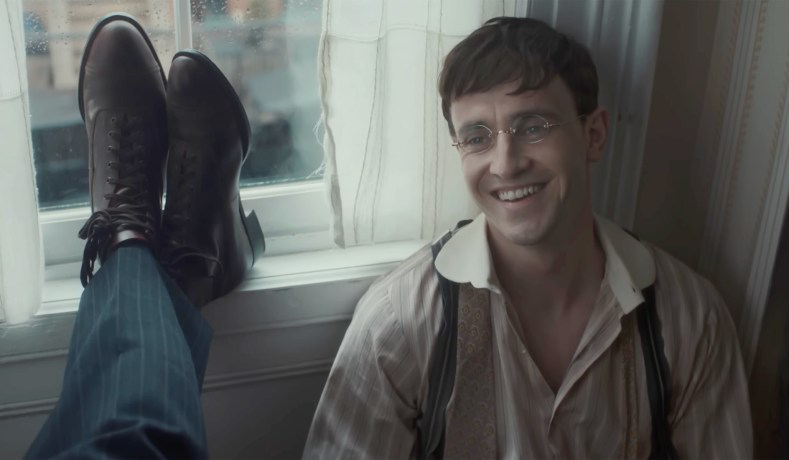 Cena do filme A História do Som. Lionel, um homem branco de cabelos castanhos, está sentado encostado na parede, ao lado de uma janela. Ele sorri e aparenta estar alegre; veste uma camisa listrada e um suspensório cinza. Os pés de outro homem, calçados com um sapato de couro marrom, estão apoiados na mesma janela.