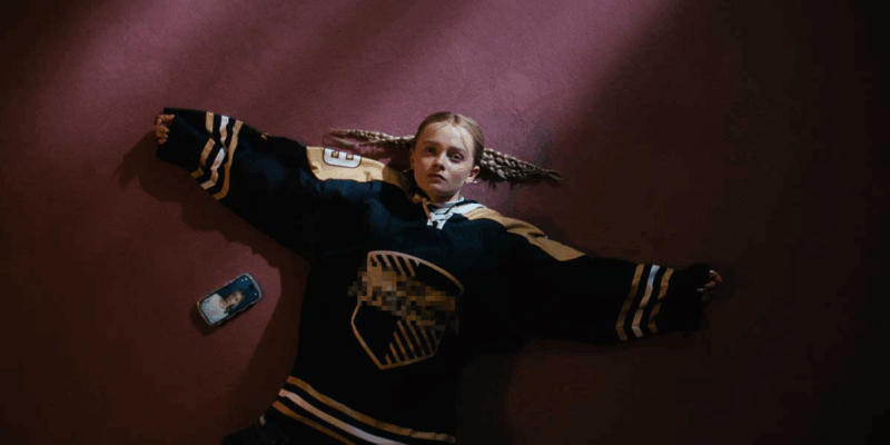  Lucas, uma menina branca de cabelos loiros aparece deitada de braços abertos em um chão vermelho. Ela veste uma camisa larga azul e amarela.