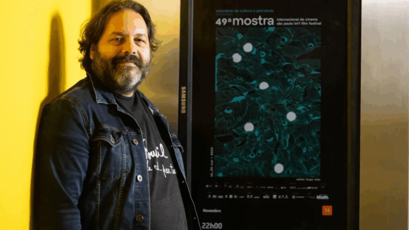 O diretor Maxime-Claude L’Écuyer, um homem de cabelos escuros e barba, vestindo uma jaqueta jeans, está em pé ao lado de um cartaz preto da 49ª Mostra Internacional de Cinema de São Paulo, contra uma parede amarela.