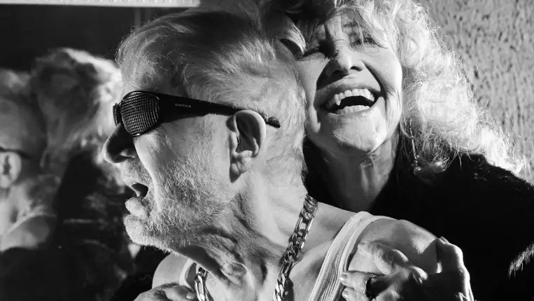 Cena do filme NosferatuNa imagem, em preto e branco, estão Helena Ignez, de vampira, e Jean-Claude Bernardet. Ela, no canto direito da foto, está abraçando o homem por trás, segurando em seu ombro com a mão esquerda. Ela está rindo, mostrando as presas dos dentes. Helena Ignez é uma mulher branca de cabelos longos, na faixa dos 80 anos. Veste uma roupa preta. Jean-Claude está de perfil, usa óculos escuros e uma corrente prateada no pescoço. Está vestindo uma regata. Ele está com a boca aberta em expressão de desespero. Jean-Claude é um homem branco, de barba e cabelos brancos, na faixa dos 85 anos. Atrás da dupla há um espelho. 