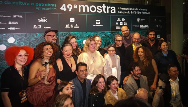 Tapete vermelho do filme Agente Secreto. Várias pessoas estão juntas enquanto olham para a câmera, para tirarem uma foto. Ao fundo está o painel da 49ª Mostra Internacional de Cinema em São Paulo.