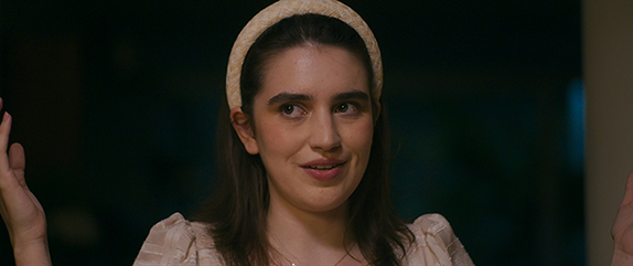 Cena do filme Virtuosas. Uma jovem mulher de cabelos castanhos escuros e lisos usa uma faixa clara na cabeça e uma blusa bege de mangas bufantes. Ela sorri levemente, com os braços erguidos e as mãos abertas, em um ambiente interno com iluminação suave