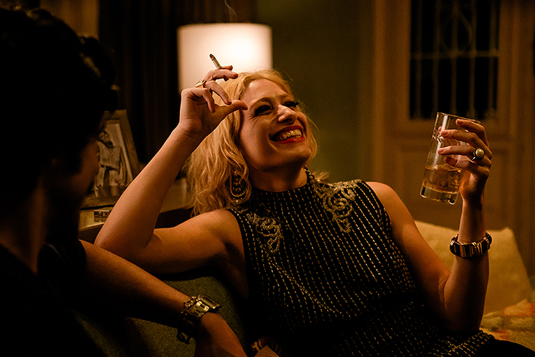 Cena do filme Homem de OuroNa imagem, a personagem Darlene está sentada em um sofá, rindo, enquanto segura um copo de vidro contendo bebida alcoólica na mão esquerda e cigarro na mão direita, que está levantada na altura de sua cabeça. Do lado esquerdo da imagem, de costas e desfocado está o personagem Mariel Mariscot, com o braço apoiado nas costas do sofá. Ele usa um relógio dourado no braço. Darlene veste uma blusa preta com várias pedras enfileiradas na vertical, que se estendem do pescoço, na gola, até a barra da roupa. Ela também usa anéis e pulseira. O cenário é uma sala de uma casa. Darlene é uma mulher branca, na faixa dos 30 anos, de cabelos loiros e lisos na altura do ombro. 