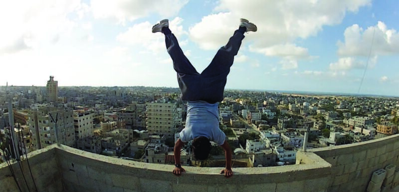 Cena do filme Yalla Parkour Na imagem, um garoto está de costas para a câmera, de ponta-cabeça na beirada de um topo de um prédio. Ele se apoia com as mãos e deixa as pernas abertas, em posição de V. No fundo, está a cidade de Gaza, com vários prédios e um céu azul com nuvens claras no horizonte. O garoto palestino veste uma camisa azul clara, tênis e calça moletom cinza. 