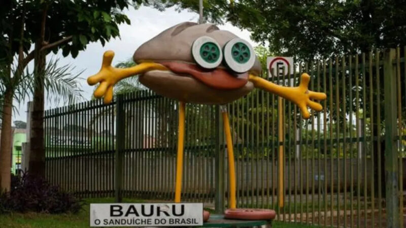 Fotografia do boneco BauruzinhoNa imagem, o boneco Bauruzinho está fixado em uma plataforma, dentro da rodoviária da cidade de Bauru. Ele é um sanduíche com olhos de pepino (na cor verde e branca), com uma carne saindo da boca, como se fosse uma língua, braços amarelos abertos, com as mãos possuindo quatro dedos, pernas amarelas e pés vermelhos, de tomate. Através dele há várias árvores e grama. Atrás, um portão de grade. Ao fundo, está a cidade, nublada. 