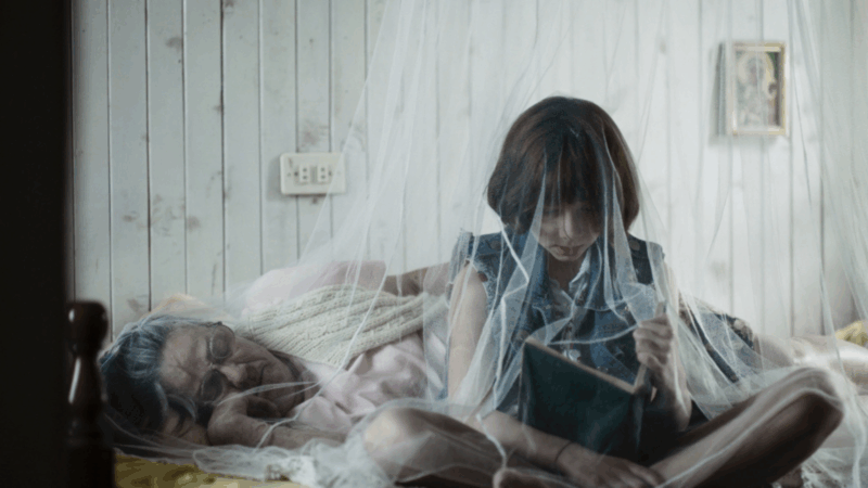 Cena do filme Delírios (2024). Uma menina lê um livro sentada na cama enquanto sua avó dorme ao lado, coberta por um mosquiteiro translúcido. A luz suave e as paredes brancas criam uma atmosfera íntima e melancólica. A rede de tule envolve as duas como se fosse uma fronteira entre o afeto e o confinamento.