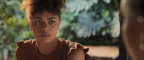 Cena do filme Criadas. Mariana, uma mulher negra, de cabelos castanhos e crespos e presos, está sentada, olhando atentamente para alguém que não aparece na imagem. Ela usa uma blusa marrom com babados nas mangas e tem expressão séria. Ao fundo, aparecem plantas verdes desfocadas.